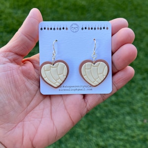 May include: A pair of heart-shaped earrings with a brown and white design. The earrings are made of polymer clay and have a gold-colored hook.