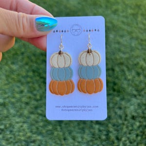 May include: A pair of dangle earrings featuring three stacked polymer clay pumpkins. The pumpkins are painted in shades of orange, blue, and white. The earrings are attached to gold hooks.
