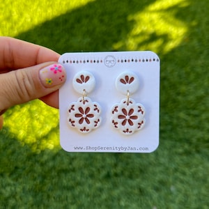 May include: A pair of white and brown floral dangle earrings. The earrings feature a small white circle with a brown flower design, and a larger white circle with a brown floral pattern.