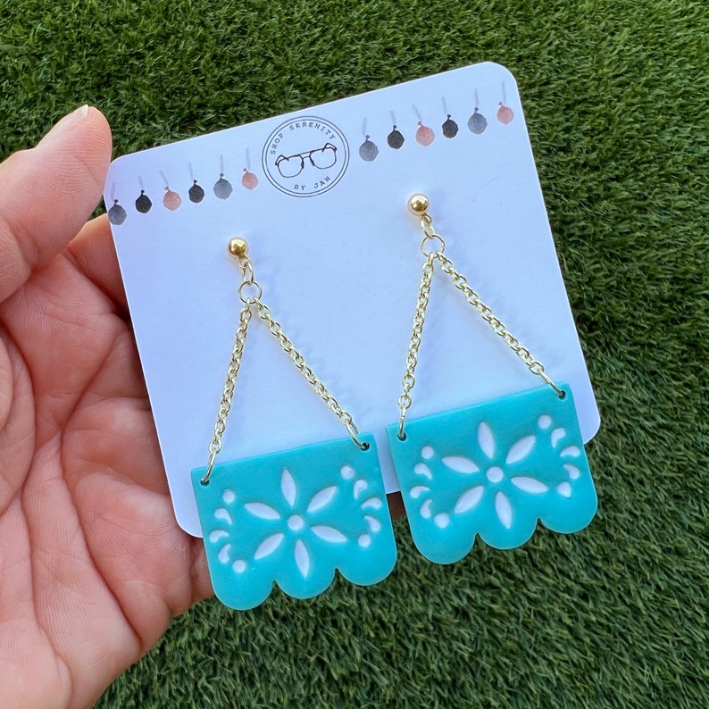 May include: Turquoise earrings with a floral design and gold-coloured chain. The earrings are on a white card with the text "SHOP SERENITY BY JAN". The earrings have a scalloped bottom edge.