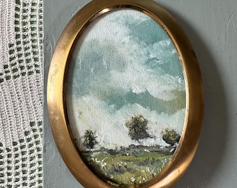 May include: A small oval painting of a landscape with a cloudy sky and green trees, framed in a gold oval frame with a stand.