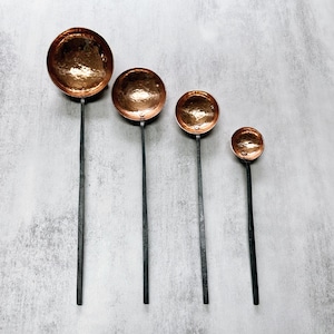 May include: Four copper ladles with black handles of varying sizes. The ladles are arranged in a row from largest to smallest.