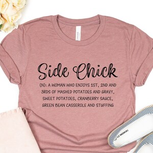 May include: A pink t-shirt with the text "Side Chick (N): A woman who enjoys 1st, 2nd and 3rds of mashed potatoes and gravy, sweet potatoes, cranberry sauce, green bean casserole and stuffing"