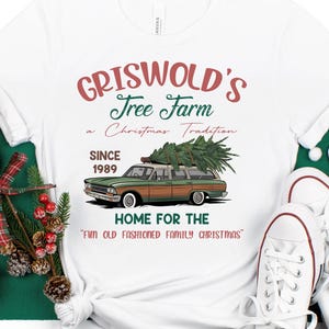 May include: White t-shirt with a graphic of a station wagon with a Christmas tree, text reads "Griswold's Tree Farm" and "Home for the Fun Old Fashioned Family Christmas." The shirt is styled with white trainers.