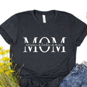 May include: A dark grey t-shirt with the word "MOM" in white letters. The word "MOM" is split in half with the names "Ariana", "Linda", and "Mina" written in cursive underneath.