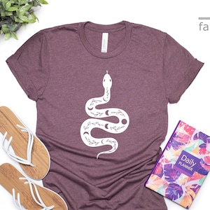 以下が含まれることがあります： 白い月と花のアクセントが付いたヘビの白いグラフィックがプリントされた、マルーンのTシャツ。Tシャツは白い表面に置かれ、白いビーチサンダルと花柄のプランナーが置かれています。