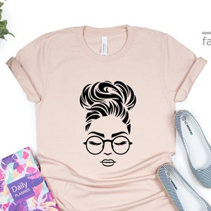 Peut inclure: Un t-shirt rose p&acirc;le avec un motif noir d'une femme avec un chignon et des lunettes. Le motif est centr&eacute; sur le t-shirt.
