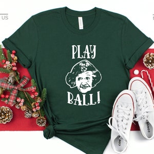 May include: A green t-shirt with a white graphic of a woman with the text "PLAY BALL!" printed on it. The t-shirt is on a red and white background with Christmas decorations.