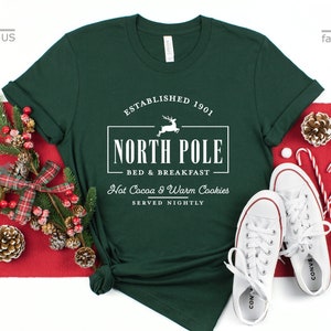May include: A forest green t-shirt with a white graphic design that says "Established 1901 North Pole Bed & Breakfast Hot Cocoa & Warm Cookies Served Nightly".
