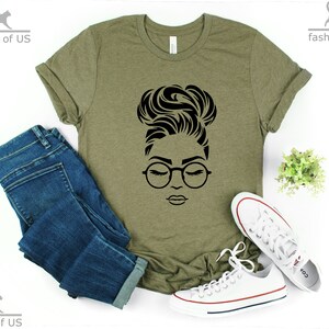 Peut inclure: Un t-shirt vert avec un motif noir repr&eacute;sentant une femme avec les cheveux en chignon et portant des lunettes. La femme regarde vers le bas, les yeux ferm&eacute;s.
