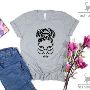 Peut inclure: Un t-shirt gris avec une illustration en noir et blanc d'une femme portant des lunettes et un chignon.