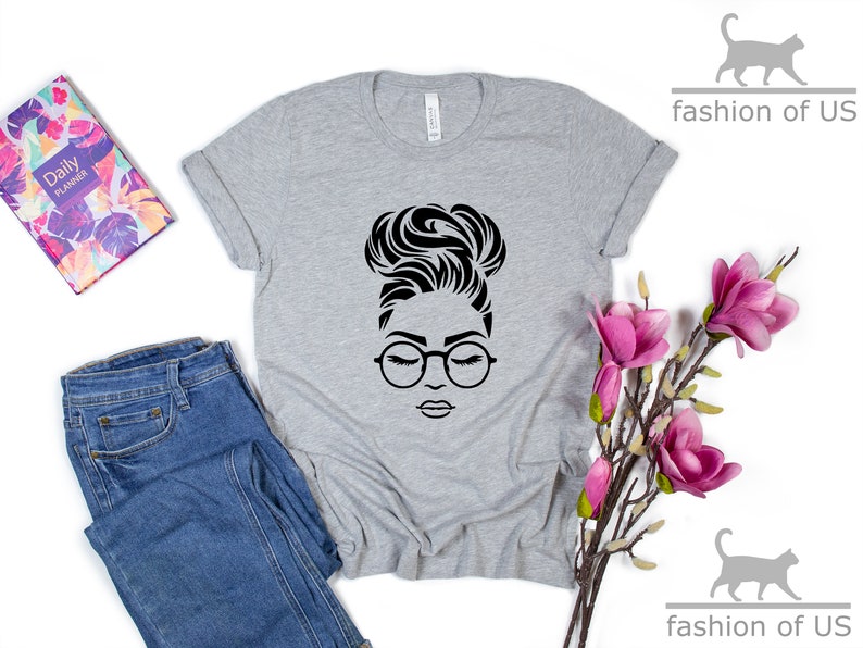 Peut inclure: Un t-shirt gris avec une illustration en noir et blanc d'une femme portant des lunettes et un chignon.