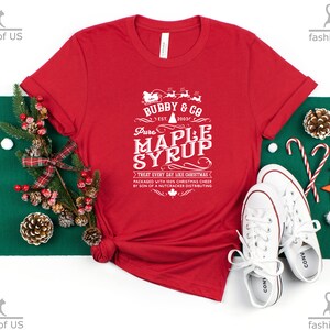 May include: Red t-shirt with a white graphic design that says "Bubby & Co. Est. 2003 Pure Maple Syrup Treat Every Day Like Christmas Packaged with 100% Christmas Cheer by Son of a Nutcracker Distributing".