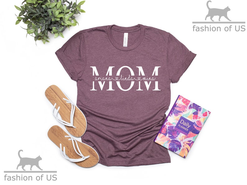 May include: A mauve t-shirt with the word "MOM" in white letters. The names "Ariana", "Linda", and "Mina" are written in a smaller font above and below the letters. A heart symbol is between the names "Linda" and "Mina".