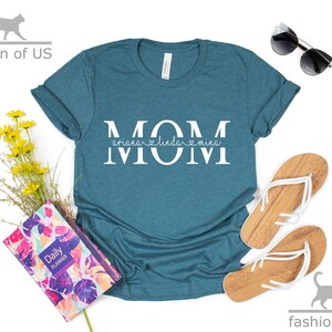 May include: Teal blue t-shirt with the word "MOM" in white letters. The word "MOM" is split in half with the names "Ariana", "Linda", and "Mina" written in smaller white letters underneath.