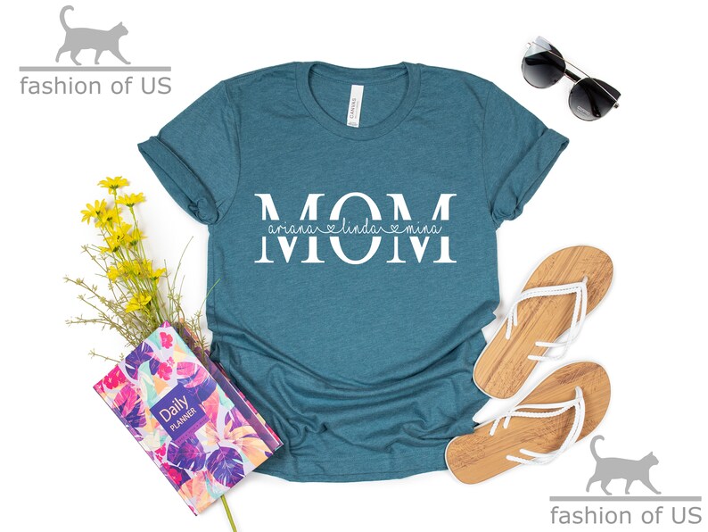 May include: Teal blue t-shirt with the word "MOM" in white letters. The word "MOM" is split in half with the names "Ariana", "Linda", and "Mina" written in smaller white letters underneath.