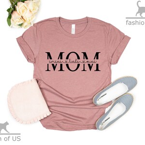 May include: A pink t-shirt with the word "MOM" in black letters. The letters are split with the names "Ariana", "Linda", and "Mina" written in a smaller font underneath.