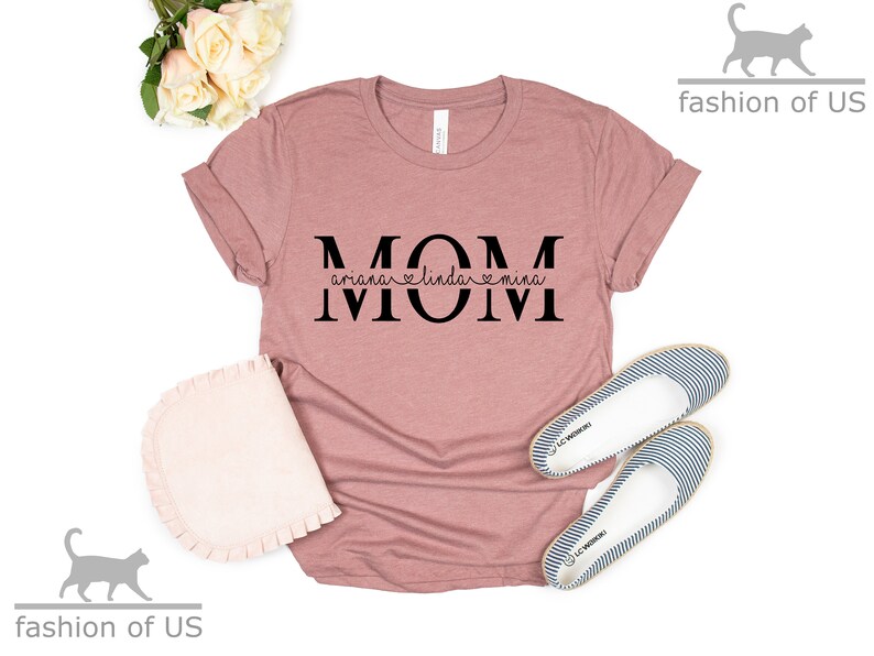May include: A pink t-shirt with the word "MOM" in black letters. The letters are split with the names "Ariana", "Linda", and "Mina" written in a smaller font underneath.