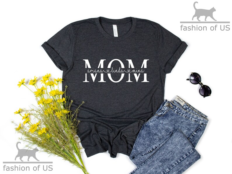 May include: A dark grey t-shirt with the word "MOM" in white letters. The word "MOM" is split in half with the names "Ariana", "Linda", and "Mina" written in cursive underneath.