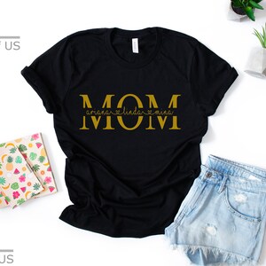 May include: Black t-shirt with the word "MOM" in gold letters. The word "MOM" is separated by hearts with the names "Ariana", "Linda", and "Mina" written underneath.