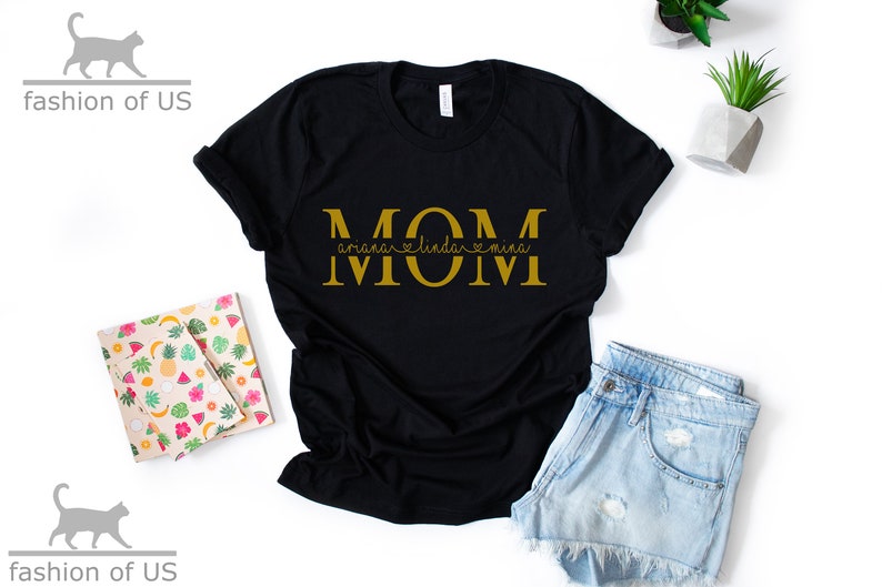 May include: Black t-shirt with the word "MOM" in gold letters. The word "MOM" is separated by hearts with the names "Ariana", "Linda", and "Mina" written underneath.