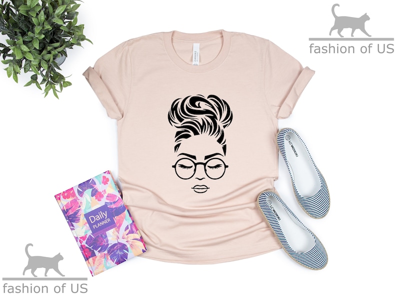 Peut inclure: Un t-shirt rose p&acirc;le avec un motif noir d'une femme avec un chignon et des lunettes. Le motif est centr&eacute; sur le t-shirt.