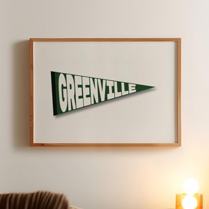 以下が含まれることがあります： 白い壁に掛けられた、緑色のペンナントに白い文字で「GREENVILLE」と書かれた額装されたプリント。寝室のインテリアです。