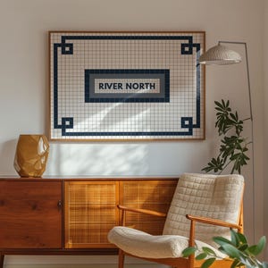 May include: A framed wall art piece with the text "RIVER NORTH" in a navy blue rectangle, set against a grid pattern. The artwork hangs above a wooden cabinet and a beige armchair. A gold-colored vase and a floor lamp are also visible.