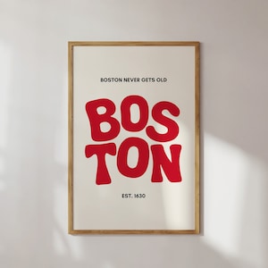 Op de afbeelding: Een ingelijste print met een witte achtergrond en zwarte tekst die "BOSTON NEVER GETS OLD" luidt. Het woord "BOSTON" staat in grote, rode, gestileerde letters. De tekst "EST. 1630" staat onder het woord "BOSTON".