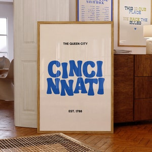 Puede incluir: Un póster blanco con un gráfico azul y blanco que dice "The Queen City" y "Cinci Nnati" con el texto "Est. 1788" debajo. El póster está enmarcado en madera y colgado en una pared.