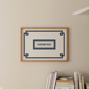 Può includere: Un'opera d'arte a mosaico incorniciata con una cornice in legno chiaro. Il mosaico presenta un motivo a griglia con un bordo blu navy e un rettangolo centrale con il testo "CUSTOM TEXT". Lo sfondo è beige chiaro.
