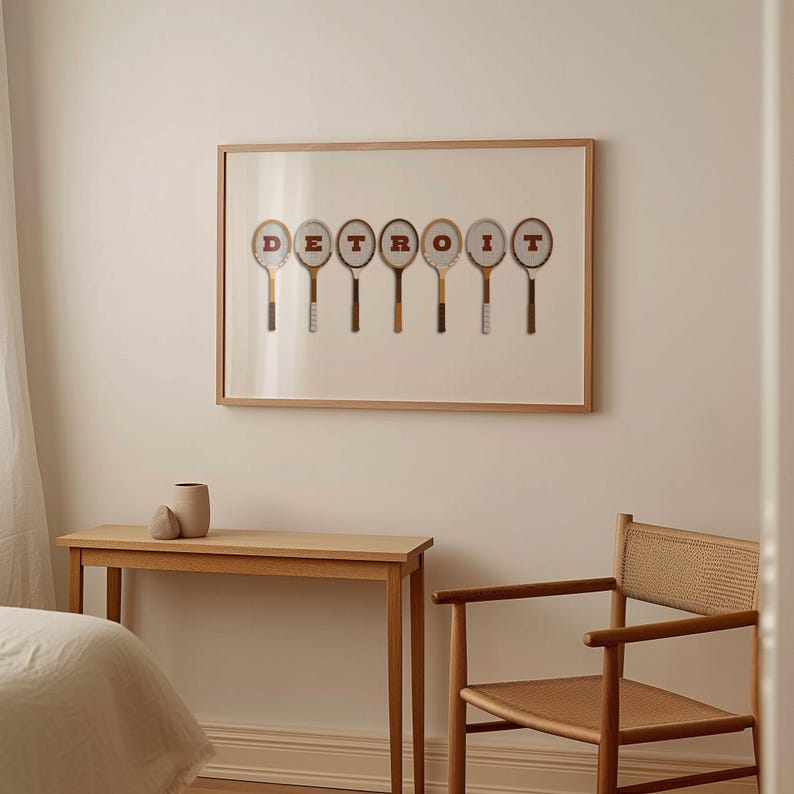 May include: A framed print of the word 'DETROIT' spelled out using vintage tennis rackets. The rackets are brown and white with red lettering. The print is hanging on a white wall above a wooden table and chair.