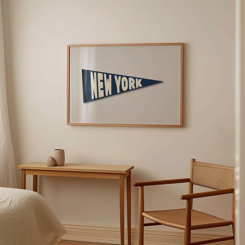 Puede incluir: Una impresi&oacute;n enmarcada de un bander&iacute;n azul con el texto "NEW YORK" en letras blancas. La impresi&oacute;n est&aacute; colgada en una pared blanca sobre una mesa y una silla de madera.