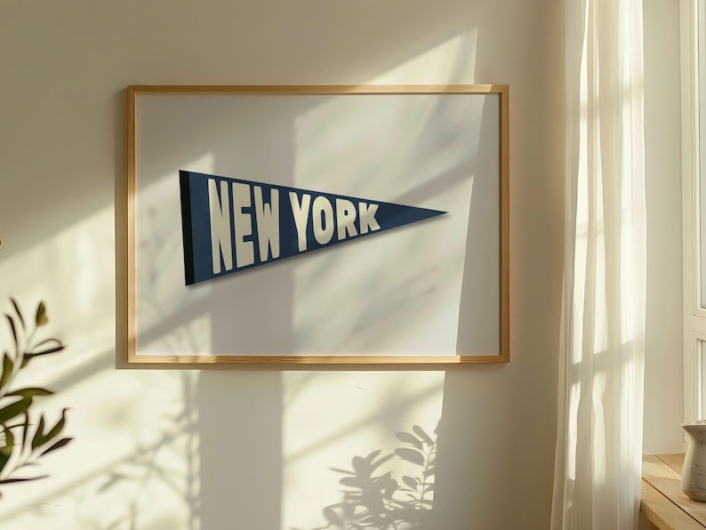 Puede incluir: Una impresi&oacute;n enmarcada de un bander&iacute;n azul con texto blanco que dice "NEW YORK". La impresi&oacute;n est&aacute; colgada en una pared blanca con un marco de madera.