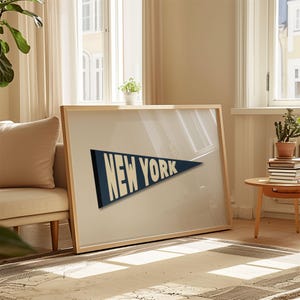 Puede incluir: Una impresi&oacute;n enmarcada de un bander&iacute;n azul con el texto "NEW YORK" en blanco. La impresi&oacute;n est&aacute; colgada en una pared en una habitaci&oacute;n con un sof&aacute; y una mesa auxiliar.