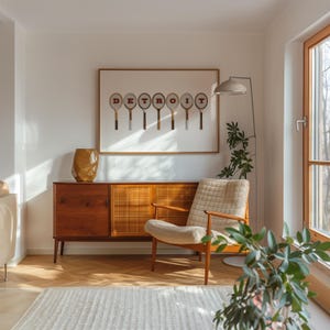 May include: A living room interior with a wooden cabinet, a white armchair, a floor lamp, and a framed print with the word 'DETROIT' spelled out using tennis rackets. The print is on a white background with a brown frame. The cabinet has two wooden doors and two woven doors. The armchair has a white fabric cushion and a wooden frame. The floor lamp has a white shade and a silver base. The floor is a light brown wood with a herringbone pattern. There is a green plant in the corner of the room.