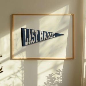 Puede incluir: Un banderín enmarcado con el texto "LAST NAME" en letras blancas sobre un fondo azul marino. El banderín está enmarcado en un marco de madera marrón claro y cuelga de una pared blanca.