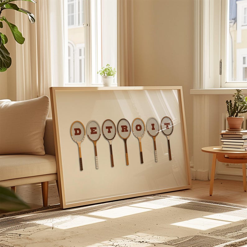 May include: A framed print with the word 'DETROIT' spelled out using seven vintage tennis rackets. The rackets are arranged horizontally and each racket has a letter of the word painted on it in red. The background of the print is white.