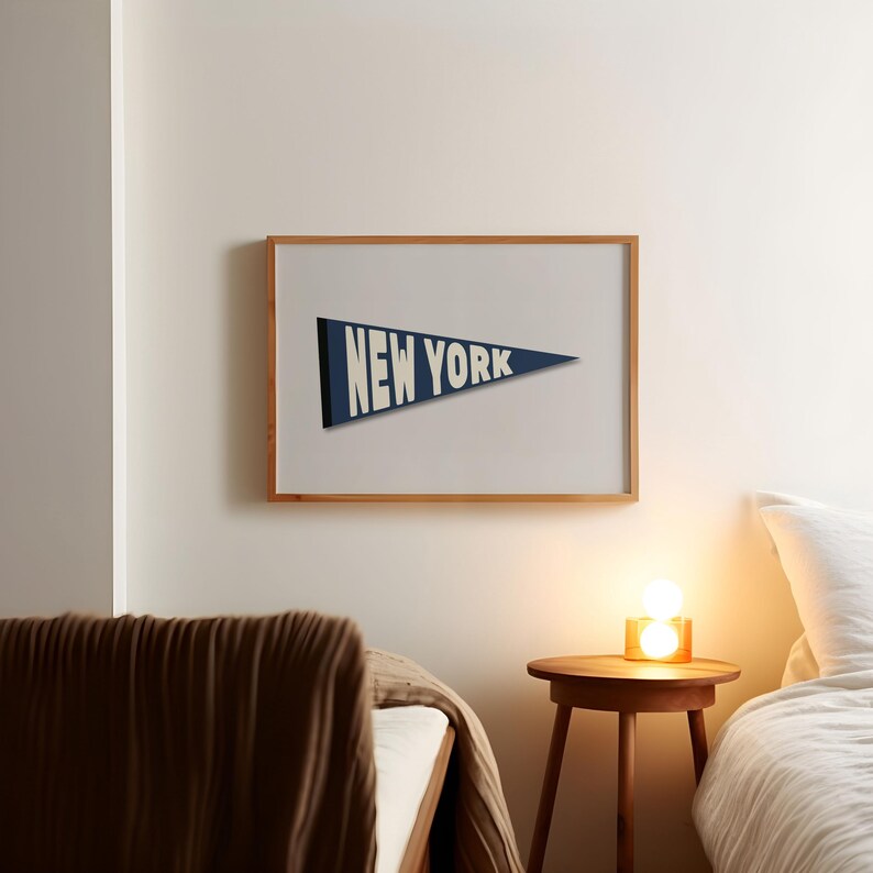 Puede incluir: Una impresi&oacute;n enmarcada de un bander&iacute;n azul con el texto "NEW YORK" en blanco. La impresi&oacute;n est&aacute; colgada en una pared blanca sobre una mesita de noche de madera con una l&aacute;mpara.