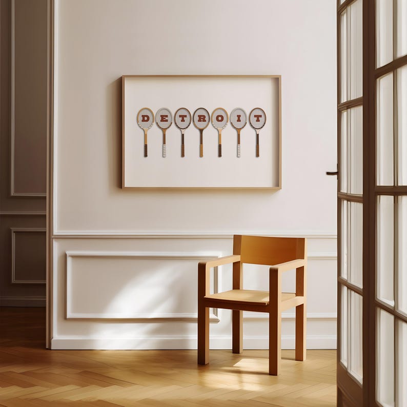 May include: A framed print of the word 'DETROIT' spelled out using vintage tennis rackets. The rackets are arranged in a row, with the letters of the word spelled out in red on the rackets. The print is hanging on a white wall, and there is a wooden chair in the foreground.