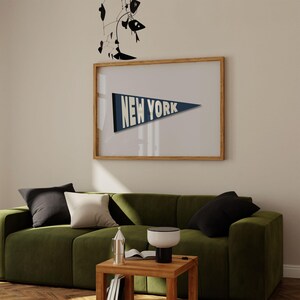 Puede incluir: Un sof&aacute; de terciopelo verde con almohadas de color blanco y negro se encuentra frente a una impresi&oacute;n enmarcada de un bander&iacute;n azul con el texto "NEW YORK" en blanco. Una peque&ntilde;a mesa de madera con una l&aacute;mpara y un libro se encuentra frente al sof&aacute;.