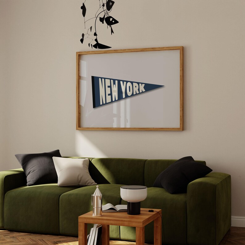 Puede incluir: Un sof&aacute; de terciopelo verde con almohadas de color blanco y negro se encuentra frente a una impresi&oacute;n enmarcada de un bander&iacute;n azul con el texto "NEW YORK" en blanco. Una peque&ntilde;a mesa de madera con una l&aacute;mpara y un libro se encuentra frente al sof&aacute;.