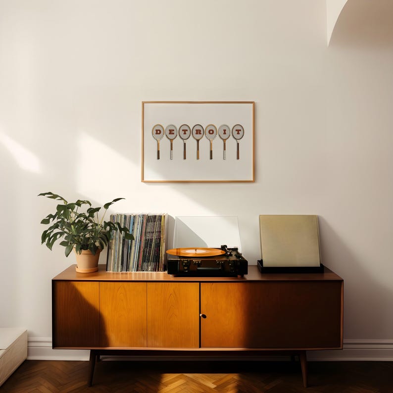May include: A mid-century modern wooden cabinet with a record player on top. The cabinet has two doors and is a light brown color. There are records stacked on the left side of the cabinet. A framed print with the word 'DETROIT' spelled out with tennis rackets is hanging on the wall behind the cabinet.