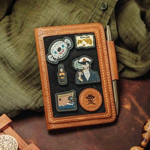 May include: A brown leather notebook with a black interior, adorned with pirate-themed patches. Patches include a skull, a pirate ship, and a skull-faced character. A pen is secured on the side. The notebook is on a dark brown surface.
