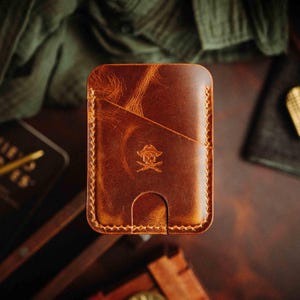 May include: A rectangular brown leather card holder featuring a diagonal pocket and a central cutout for easy card access. The surface displays a distressed texture and a stamped emblem of a skull with crossed swords centred on the front panel.