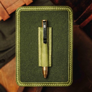 Puede incluir: Un bolígrafo con punta de latón y un clip negro con diseño de calavera, alojado en un estuche de cuero verde. El bolígrafo y el estuche están dentro de un estuche de cuero verde más grande con bordes cosidos.