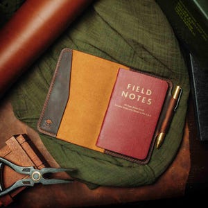 May include: A brown leather notebook cover with a tan interior and a red Field Notes notebook inside. The notebook cover has a pen holder and a skull and crossbones logo.