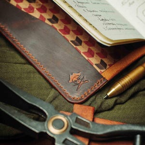 May include: A brown leather notebook cover with a red and white geometric pattern on the inside. The cover has a stitched edge and a skull and crossbones design embossed on the front.
