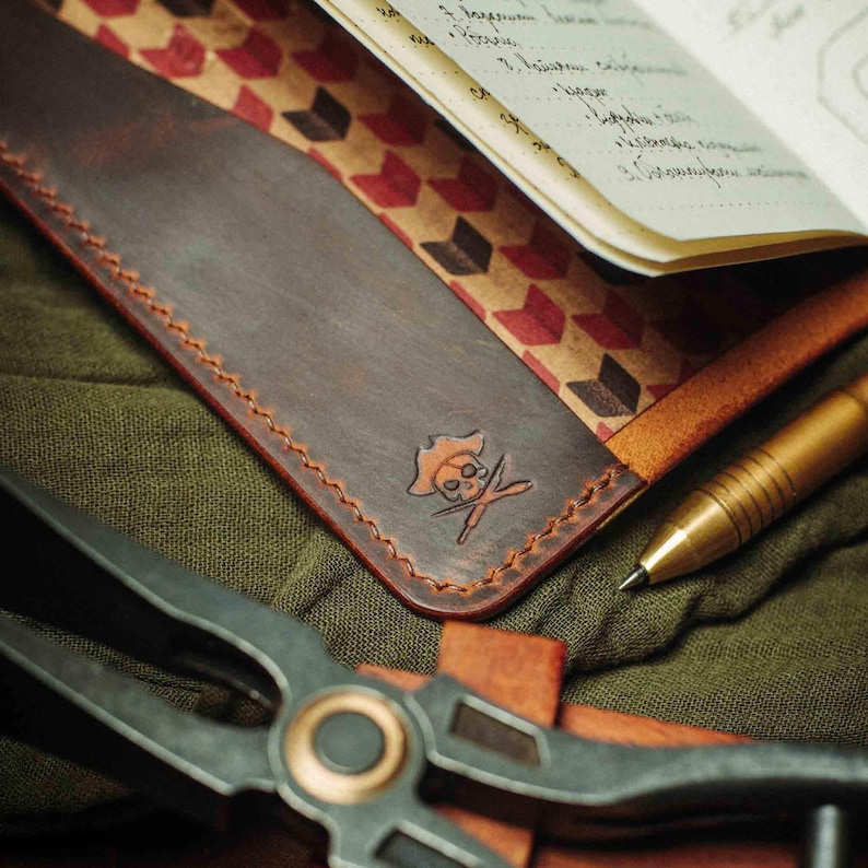 May include: A brown leather notebook cover with a red and white geometric pattern on the inside. The cover has a stitched edge and a skull and crossbones design embossed on the front.