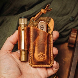 Può includere: Un astuccio in pelle marrone con una penna in ottone e un coltello svizzero. L'astuccio presenta un logo pirata con teschio e ossa incrociate. La penna ha una clip dorata. Il coltello ha un manico in legno.